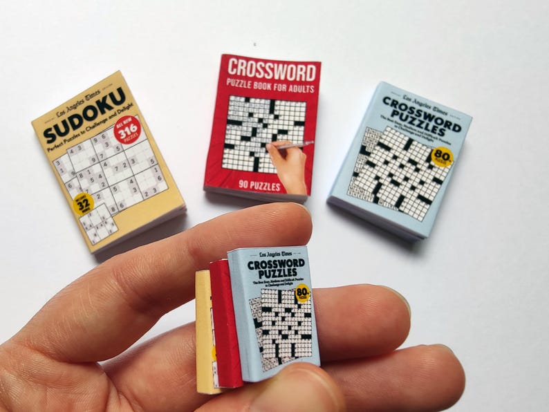 K&ouml;nnte beinhalten: Eine Hand h&auml;lt Miniatur-Puzzleb&uuml;cher, darunter Sudoku- und Kreuzwortr&auml;tsel. Die B&uuml;cher haben farbenfrohe Einb&auml;nde mit Texten wie "Los Angeles Times" und "Crossword Puzzles". Das Sudoku-Buch enth&auml;lt 316 R&auml;tsel.