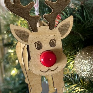 May include: A wooden reindeer ornament with a red bottle cap nose. The reindeer has brown antlers and a light brown body. The ornament is hanging on a Christmas tree.