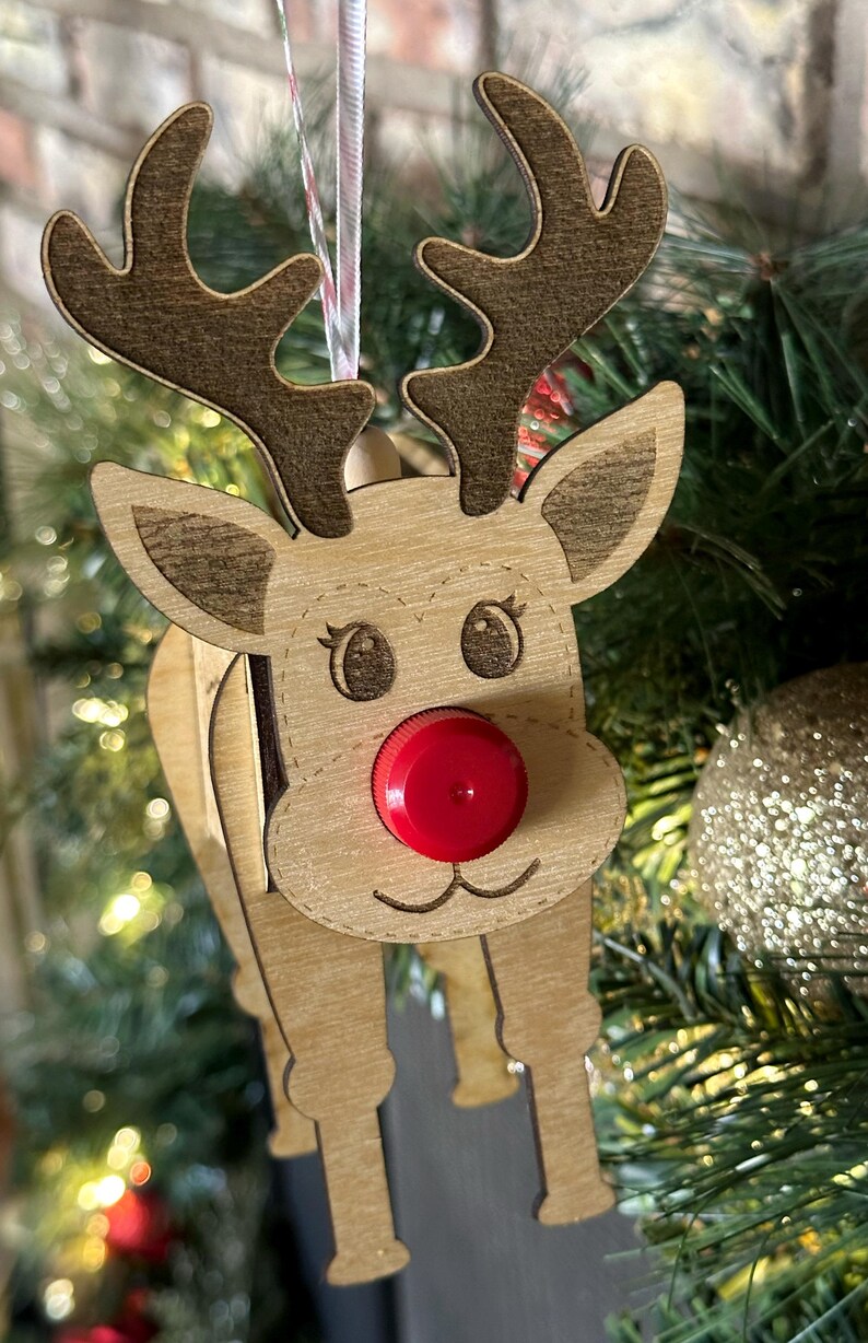 May include: A wooden reindeer ornament with a red bottle cap nose. The reindeer has brown antlers and a light brown body. The ornament is hanging on a Christmas tree.