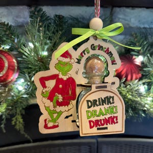 May include: A wooden Christmas ornament featuring the Grinch in a red Santa suit and a small bottle with the text "Drink! Drank! Drunk!" The ornament is decorated with green and red accents and has the text "Merry Grinchmas!"