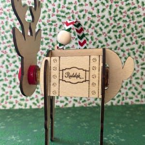 May include: Wooden reindeer ornament with a red nose, a red and white striped ribbon, and the name "Rudolph" written on the side. The ornament is standing on a green background with a snowflake pattern.