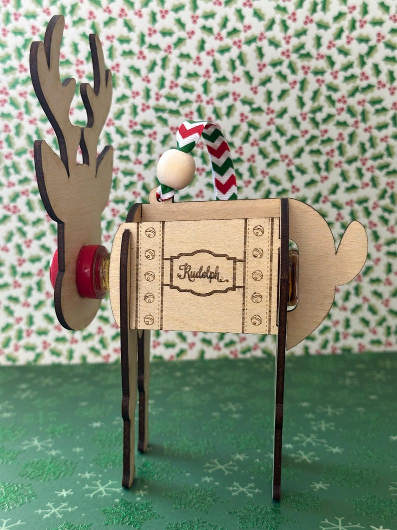 May include: Wooden reindeer ornament with a red nose, a red and white striped ribbon, and the name "Rudolph" written on the side. The ornament is standing on a green background with a snowflake pattern.