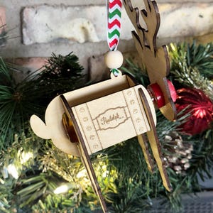 May include: Wooden reindeer Christmas ornament with a red and green striped ribbon, a wooden bead, and the word "Rudolph" engraved on the side.