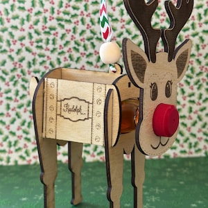 May include: Wooden reindeer ornament with a small bottle holder. The reindeer is brown and has a red nose. The bottle holder is labeled "Rudolph".