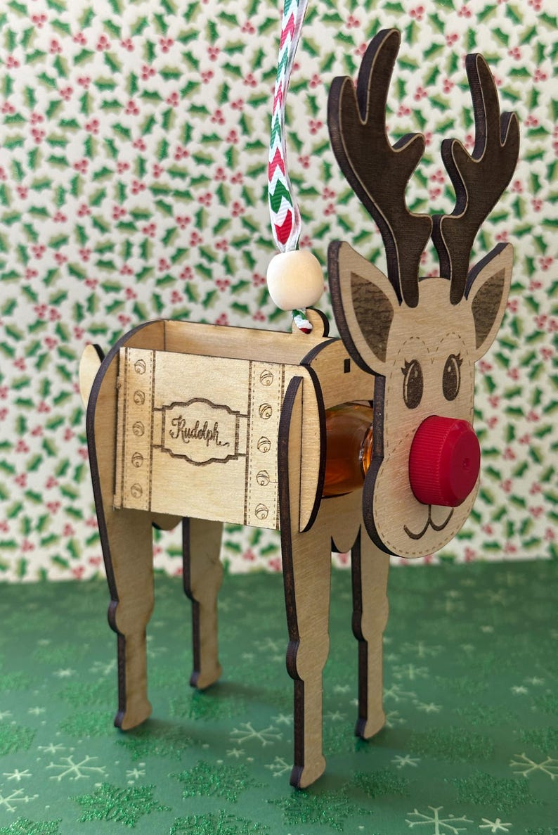 May include: Wooden reindeer ornament with a small bottle holder. The reindeer is brown and has a red nose. The bottle holder is labeled "Rudolph".