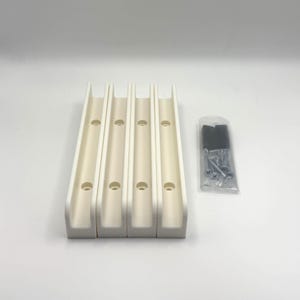 Puede incluir: Cuatro soportes de montaje de plástico en forma de U de color blanco roto, cada uno con dos orificios circulares pretaladrados para la instalación. Junto a los soportes hay una pequeña bolsa de plástico transparente con tornillos metálicos.