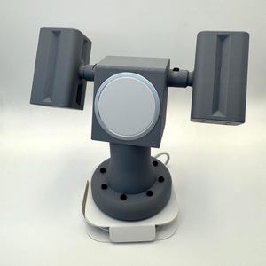 May include: A gray, 3D-printed device with two rectangular, angled components and a central circular element. The device is mounted on a white base with a curved design. The overall structure suggests a modern, tech-related product.
