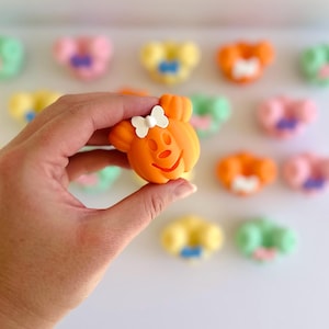 May include: An orange, cartoon-like character with a white bow, held in a hand. Other colorful, similar characters are blurred in the background. The character has a smiling face and mouse-like ears.