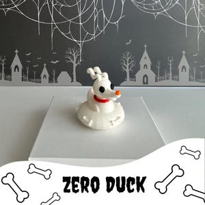 May include: A white Zero Duck figurine with a red collar and orange nose, inspired by the movie "The Nightmare Before Christmas." The figurine is on a white surface, with a dark gray background featuring a spooky Halloween scene and the text "ZERO DUCK."