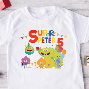 May include: White t-shirt with a colorful graphic of cartoon monsters celebrating a birthday. The text "Super Peter 5" is printed on the shirt.