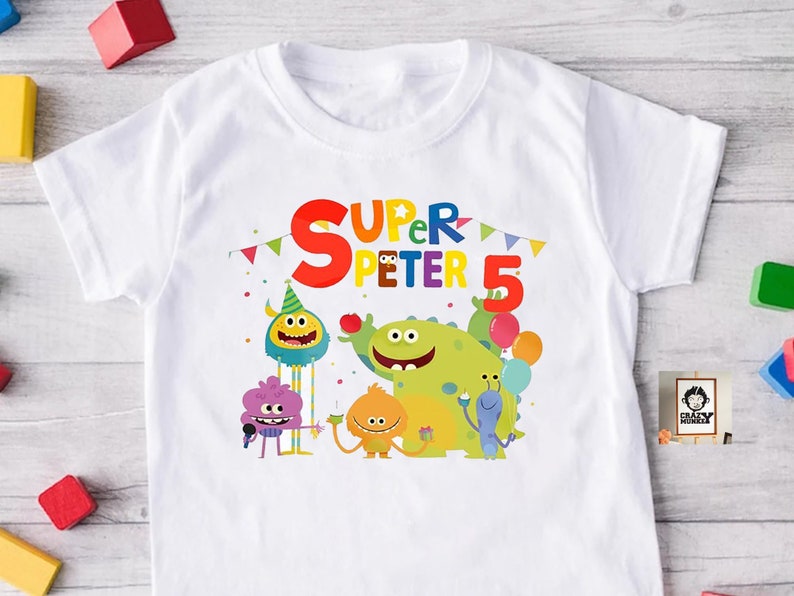 May include: White t-shirt with a colorful graphic of cartoon monsters celebrating a birthday. The text "Super Peter 5" is printed on the shirt.