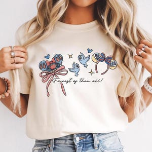 May include: A cream-coloured t-shirt with a cartoon design. The design features a bow, birds, and mouse ear headbands. The text "Fairest of them all!" is included in a script font. The graphic is in shades of blue, pink, and yellow.