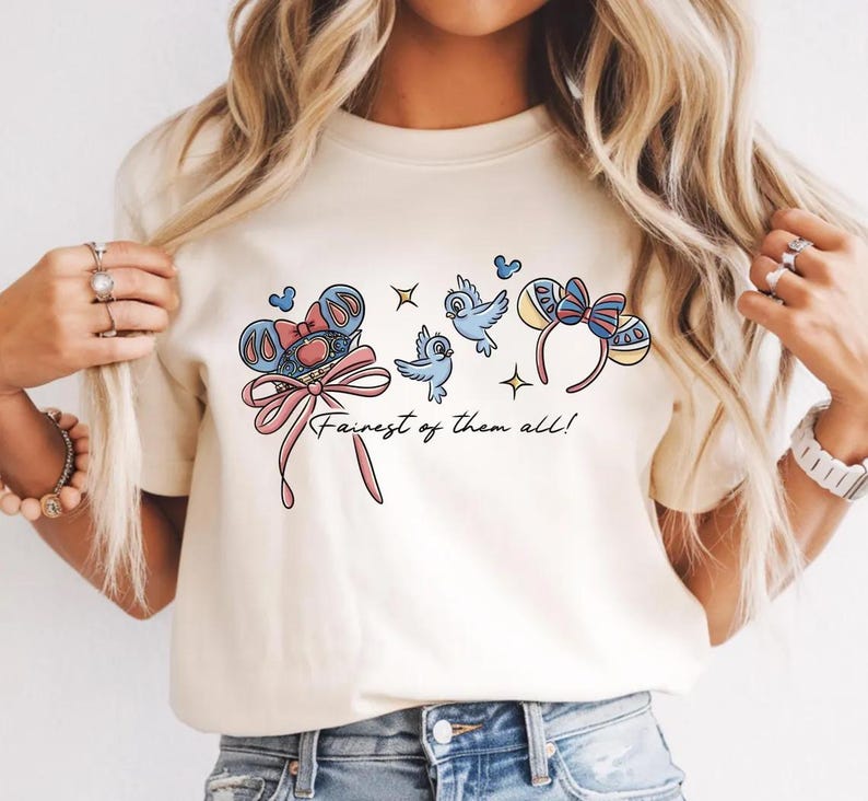 May include: A cream-coloured t-shirt with a cartoon design. The design features a bow, birds, and mouse ear headbands. The text "Fairest of them all!" is included in a script font. The graphic is in shades of blue, pink, and yellow.