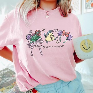 May include: Pale pink t-shirt with a mermaid-themed graphic. The design includes a seashell, a yellow fish, and purple ears with a bow, along with the text "Part of your world."