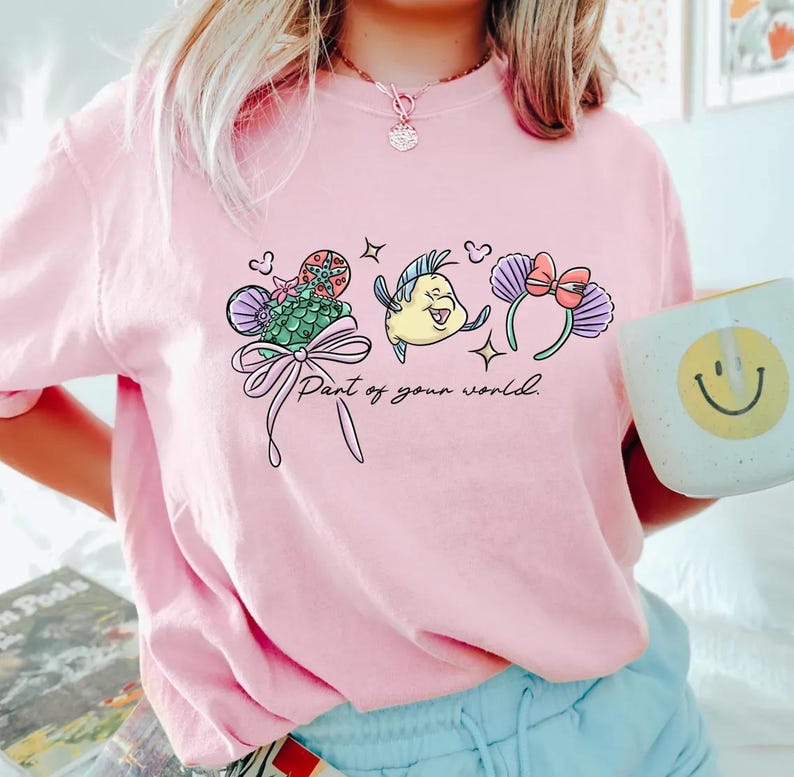 May include: Pale pink t-shirt with a mermaid-themed graphic. The design includes a seashell, a yellow fish, and purple ears with a bow, along with the text "Part of your world."