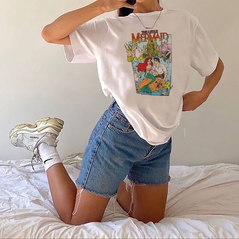 May include: A white t-shirt with a graphic print of the Disney movie "The Little Mermaid". The graphic features Ariel, Prince Eric, and other characters from the movie. The shirt is being worn by a person with blue denim shorts.