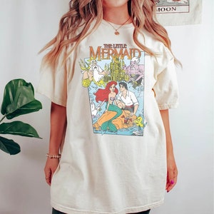 May include: A cream colored t-shirt with a graphic print of the Disney movie "The Little Mermaid". The graphic features Ariel, Prince Eric, King Triton, Ursula, and Flounder.