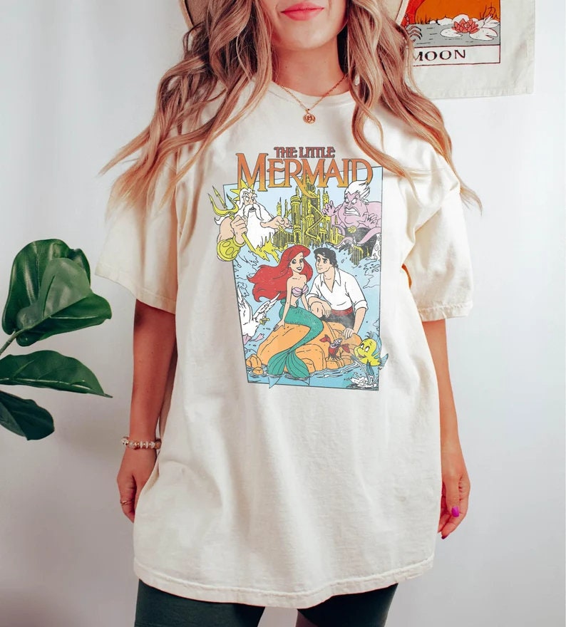 May include: A cream colored t-shirt with a graphic print of the Disney movie "The Little Mermaid". The graphic features Ariel, Prince Eric, King Triton, Ursula, and Flounder.