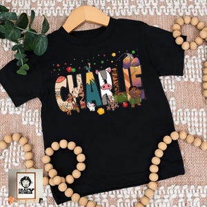 May include: Black toddler t-shirt with the name "Charlie" in colorful, patterned letters. The letters are decorated with cartoon characters and designs. The shirt is hanging on a wooden hanger.