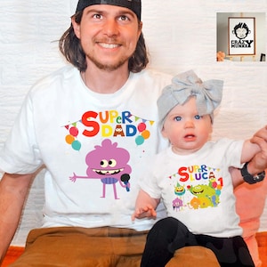 May include: White t-shirts with a colorful graphic of a cartoon character. The adult shirt says "Super Dad" and the child shirt says "Super Luca 1".