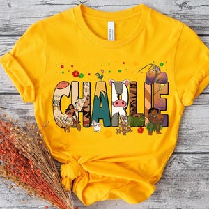 May include: Mustard yellow t-shirt with the name "CHARLIE" in colorful, illustrated letters. The letters feature characters and designs from a popular animated movie. The shirt has short sleeves and a crew neck.