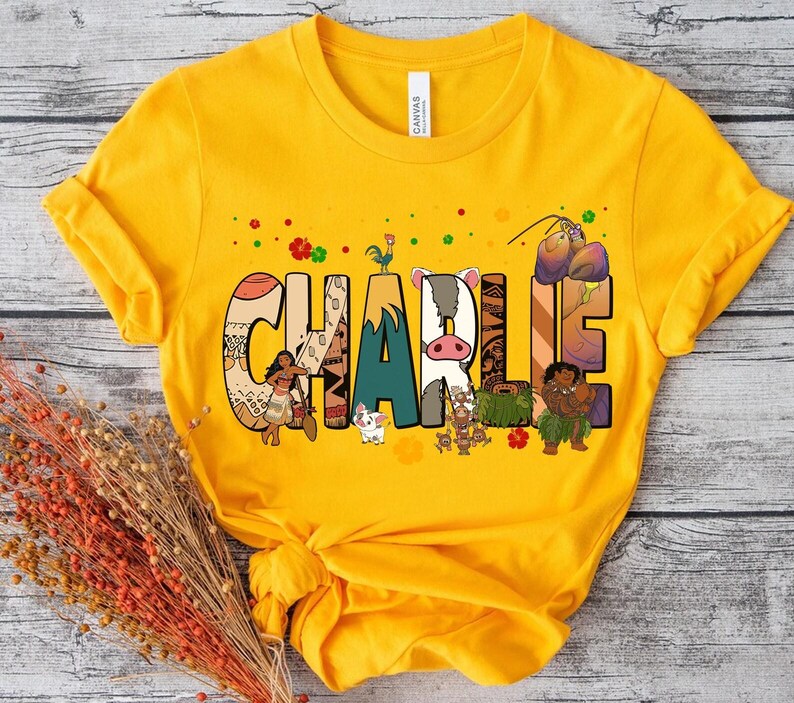 May include: Mustard yellow t-shirt with the name "CHARLIE" in colorful, illustrated letters. The letters feature characters and designs from a popular animated movie. The shirt has short sleeves and a crew neck.