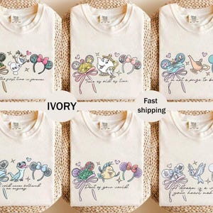 May include: Ivory t-shirts with Disney character designs. Each shirt has a different graphic featuring characters, bows, and phrases such as "Tale as old as time" and "Part of your world". The shirts are arranged on a woven surface.