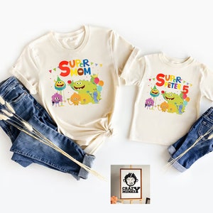 May include: Two cream colored t-shirts with a colorful graphic of cartoon characters and the text "Super Mom" and "Super Peter 5". A small wooden sign with the text "Crazy Munkee" is in the bottom right corner.