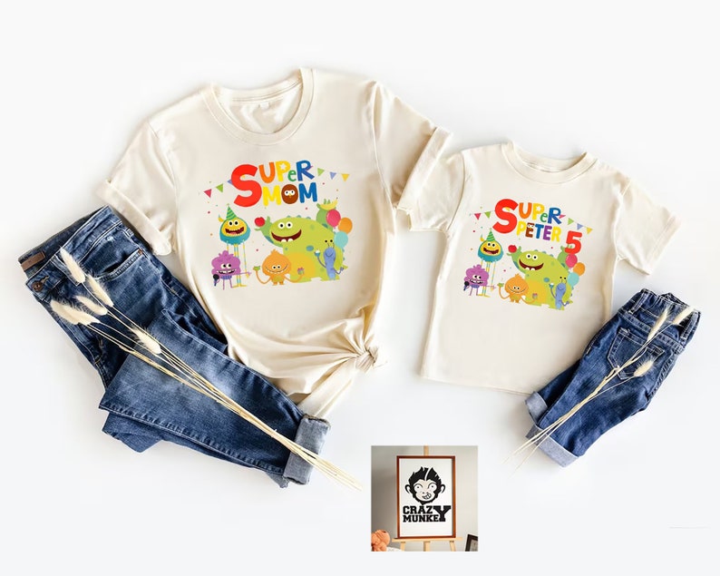 May include: Two cream colored t-shirts with a colorful graphic of cartoon characters and the text "Super Mom" and "Super Peter 5". A small wooden sign with the text "Crazy Munkee" is in the bottom right corner.