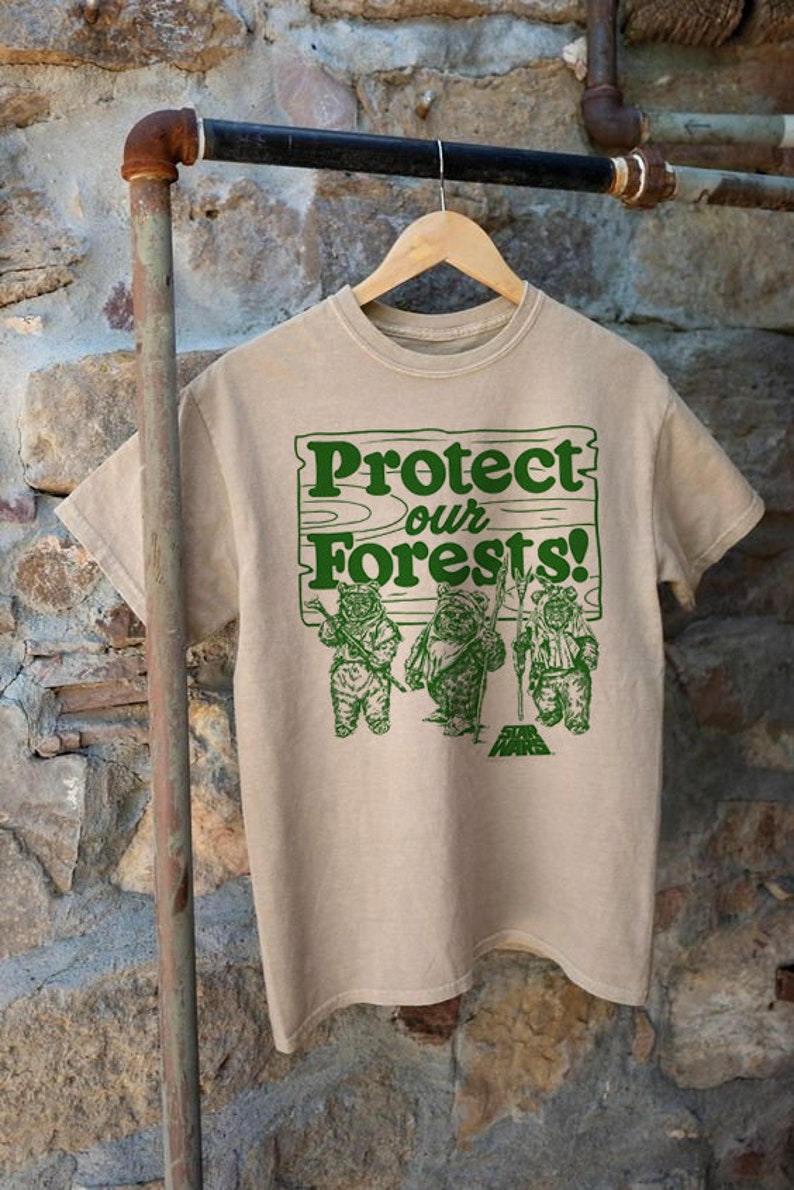 May include: A beige t-shirt with a green graphic print of three Ewoks holding signs that say "Protect our Forests!" The graphic also includes the Star Wars logo.