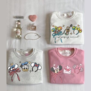 May include: Four sweatshirts arranged on a flat surface. A white sweatshirt features Mickey Mouse, Goofy, and Pluto with the text "Just goofing around!". A grey sweatshirt has Donald and Daisy Duck. A pink sweatshirt features Minnie Mouse. Accessories include a perfume bottle, a heart-shaped compact, and a bottle.