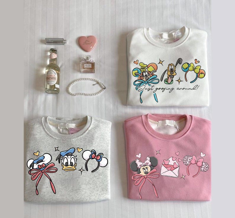 May include: Four sweatshirts arranged on a flat surface. A white sweatshirt features Mickey Mouse, Goofy, and Pluto with the text "Just goofing around!". A grey sweatshirt has Donald and Daisy Duck. A pink sweatshirt features Minnie Mouse. Accessories include a perfume bottle, a heart-shaped compact, and a bottle.