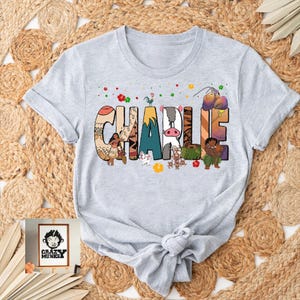 May include: A light gray t-shirt featuring the name "CHARLIE" in colorful, decorative letters. Each letter is designed with a different pattern and imagery, including cartoon characters and floral accents. The shirt has short sleeves and a crew neck.