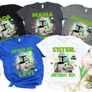 May include: A collection of t-shirts with a Star Wars theme. The shirts are black, gray, blue, and white, each featuring a graphic of The Mandalorian and Grogu. Text includes "DADA", "MAMA", "BROTHER", "BIRTHDAY BOY", and "SISTER".
