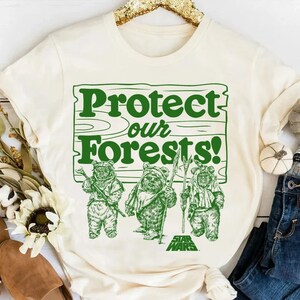 May include: A white t-shirt with a green graphic of three Ewoks from Star Wars standing in front of a sign that says "Protect our Forests!"