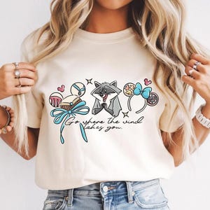 May include: A cream-coloured t-shirt with a raccoon graphic and the text "Go where the wind takes you." The design incorporates cartoon elements such as a bow, hearts, and a headband.