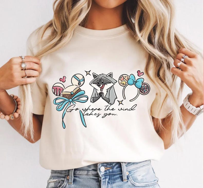 May include: A cream-coloured t-shirt with a raccoon graphic and the text "Go where the wind takes you." The design incorporates cartoon elements such as a bow, hearts, and a headband.