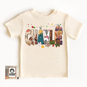 May include: Cream-colored t-shirt with the name "CHARLIE" in colorful, illustrated letters. The letters feature images of cartoon characters and tropical elements. A small framed image with a logo is in the lower left corner.