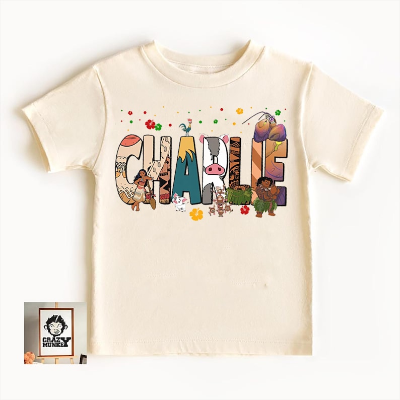 May include: Cream-colored t-shirt with the name "CHARLIE" in colorful, illustrated letters. The letters feature images of cartoon characters and tropical elements. A small framed image with a logo is in the lower left corner.