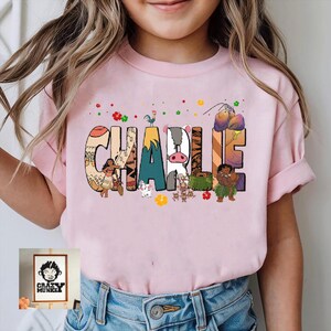May include: Pink t-shirt with the name "Charlie" in colorful, patterned letters. The letters are decorated with cartoon characters and tropical elements. The shirt is worn by a child.