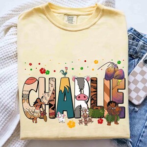 May include: A light yellow t-shirt with the name "CHARLIE" in large, colorful letters. Each letter is decorated with images of characters and elements from a popular animated movie. The shirt is made of a soft material.