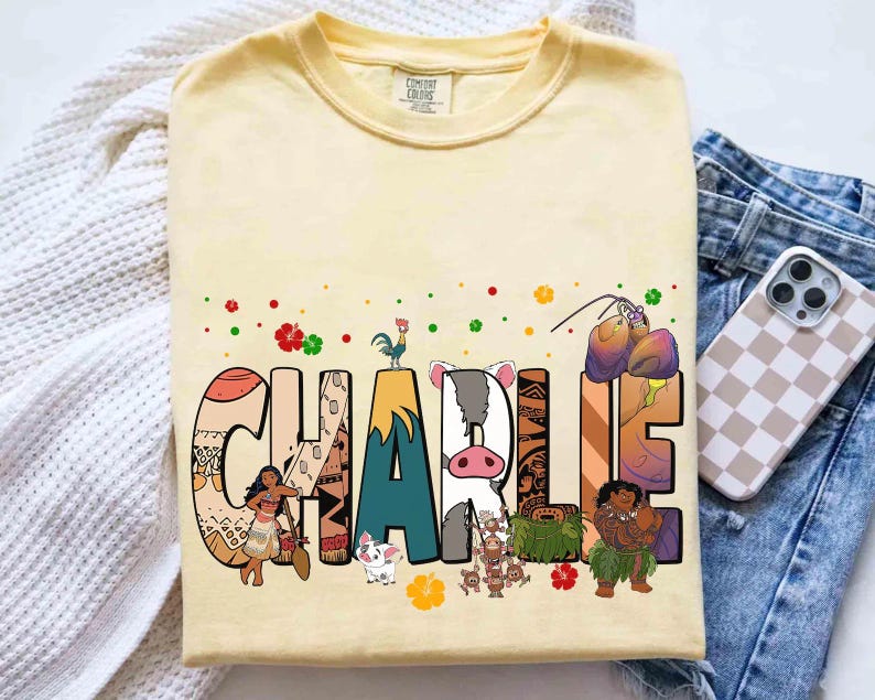 May include: A light yellow t-shirt with the name "CHARLIE" in large, colorful letters. Each letter is decorated with images of characters and elements from a popular animated movie. The shirt is made of a soft material.