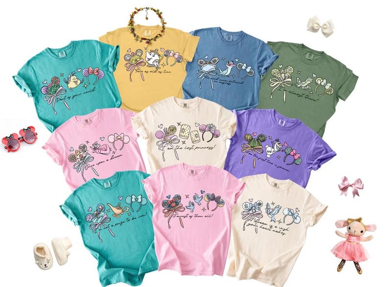 May include: A selection of colourful t-shirts featuring cartoon illustrations and text. The shirts are in shades of teal, yellow, blue, green, pink, white, and purple. Each t-shirt has a unique design with cartoon characters and bows. Accessories like sunglasses and a doll are also present.