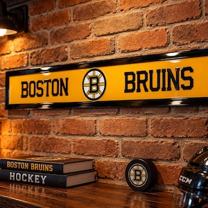 Puede incluir: Un letrero de pared rectangular de los Boston Bruins con fondo amarillo, letras negras y el logotipo del equipo en el centro, enmarcado en negro. El letrero presenta el texto BOSTON BRUINS con el logotipo de la letra B entre las palabras.