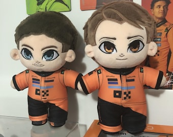May include: Two plush dolls of a person wearing an orange and black racing suit. The dolls have large eyes and brown hair. The racing suits have blue and black accents. The dolls are holding hands.