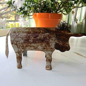 May include: A rusty metal sculpture of a cow with a white and brown patina. The cow is standing on a white surface.
