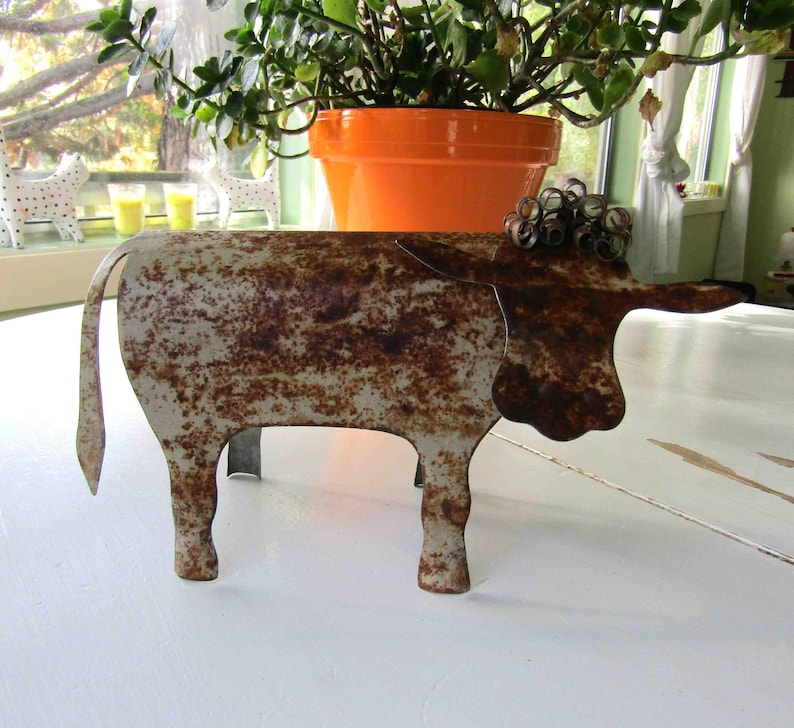 May include: A rusty metal sculpture of a cow with a white and brown patina. The cow is standing on a white surface.
