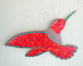 May include: A red hummingbird wall art sculpture with a turquoise outline. The hummingbird is in flight with its wings spread out. The sculpture is made of wood and has a textured surface.
