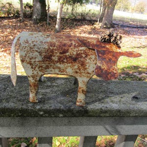 May include: A rusty metal cow sculpture with a white paint finish. The cow has a curled metal design on its head and a long tail.