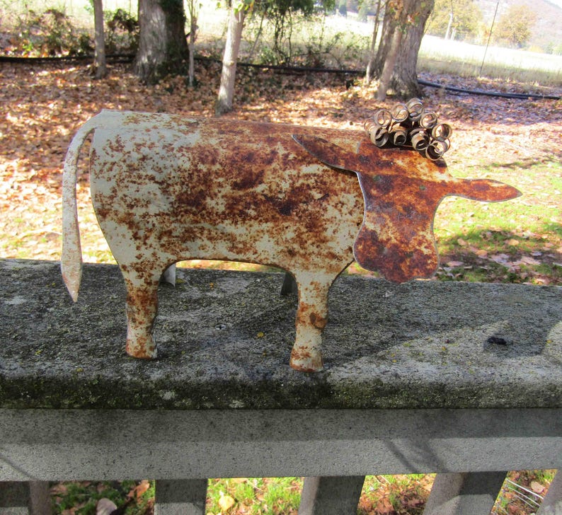 May include: A rusty metal cow sculpture with a white paint finish. The cow has a curled metal design on its head and a long tail.